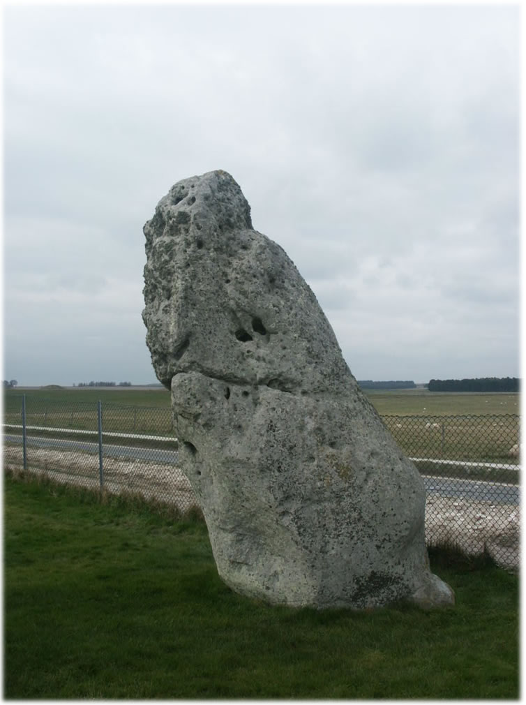 Closeup of the Heelstone, a large standing stone at Stonehenge, England. The stone is rectangular in shape and has a rough surface texture with visible cracks and lines. Its base is partially buried in the ground, and it stands tall against a cloudy sky.